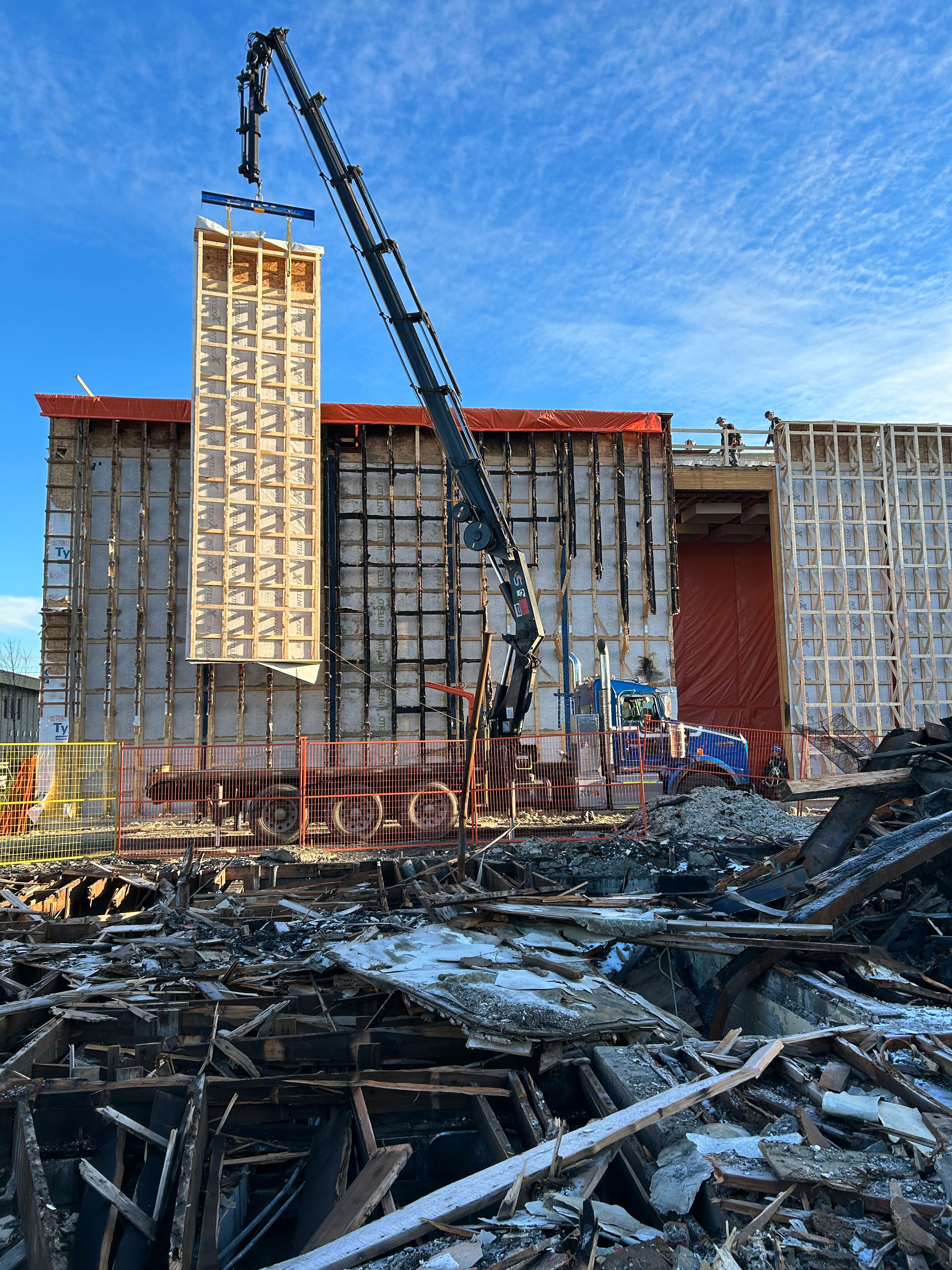 Lifting a new prefabricated wall panel into place. The damaged panels are behind, which were replaced one at a time. Note that the interior air/vapor membrane (Intello) in the damaged trusses is still intact – this is because of the mineral fiber insulation. The debris in the foreground is the building that exploded.